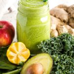 Green smoothie in a mason jar surrounded by greens, lemon, apple, ginger and avocado.
