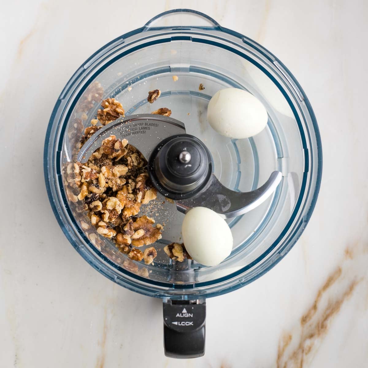 Bowl of food processor with some whole walnuts and two peeled boiled eggs.