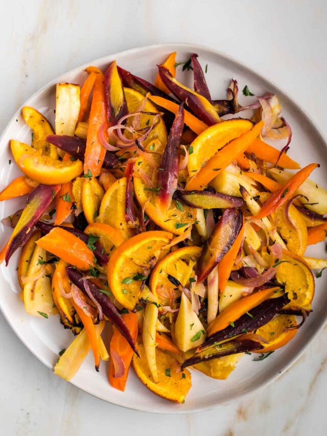 Brightly colored roasted root vegetables, purple and orange carrots, onions, parsnips and oranges.
