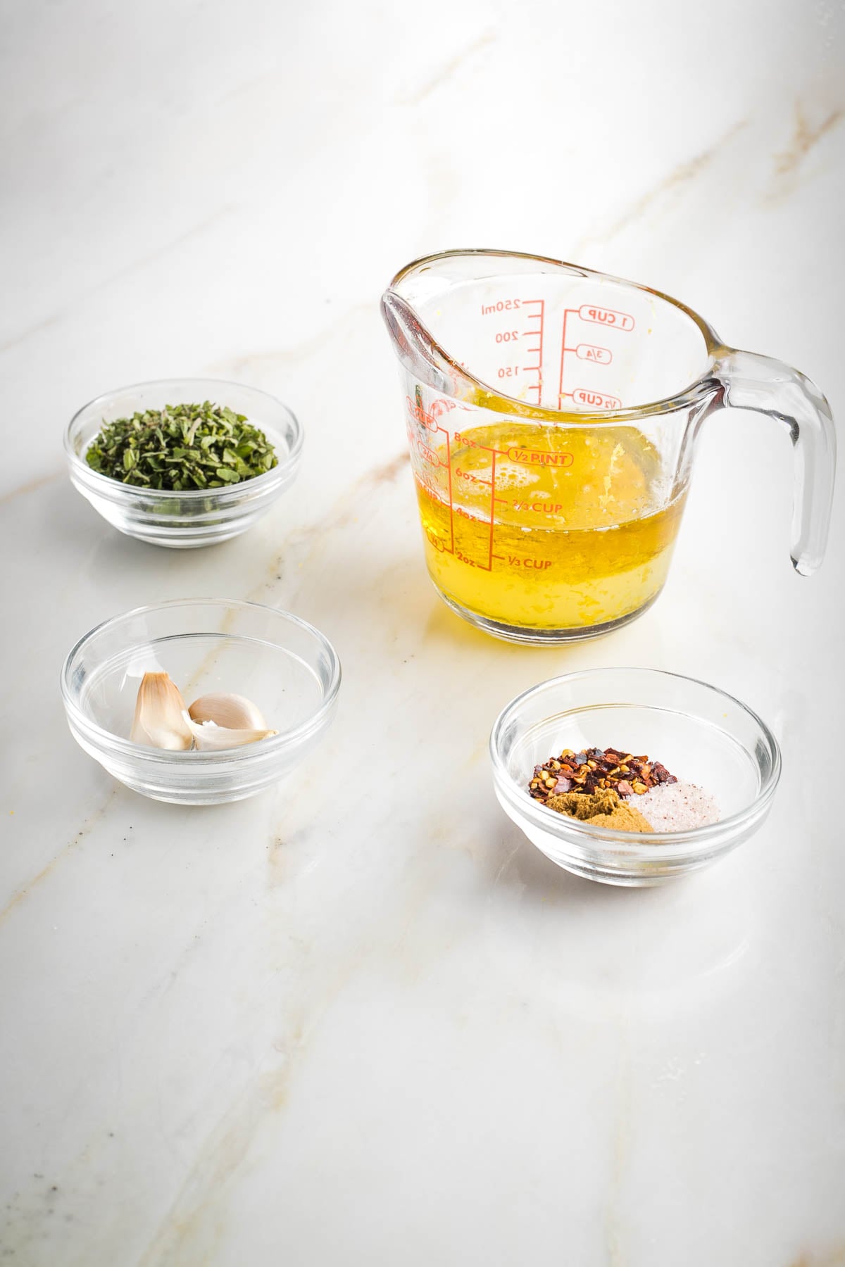 Olive oil and lemon juice in a cup, and small dishes with fresh herbs, garlic cloves and dried spices. 