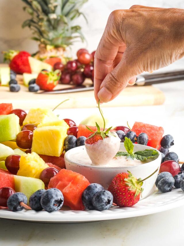 Dipping a strawberry into a creamy dip on a plate of assorted fruit kebabs.