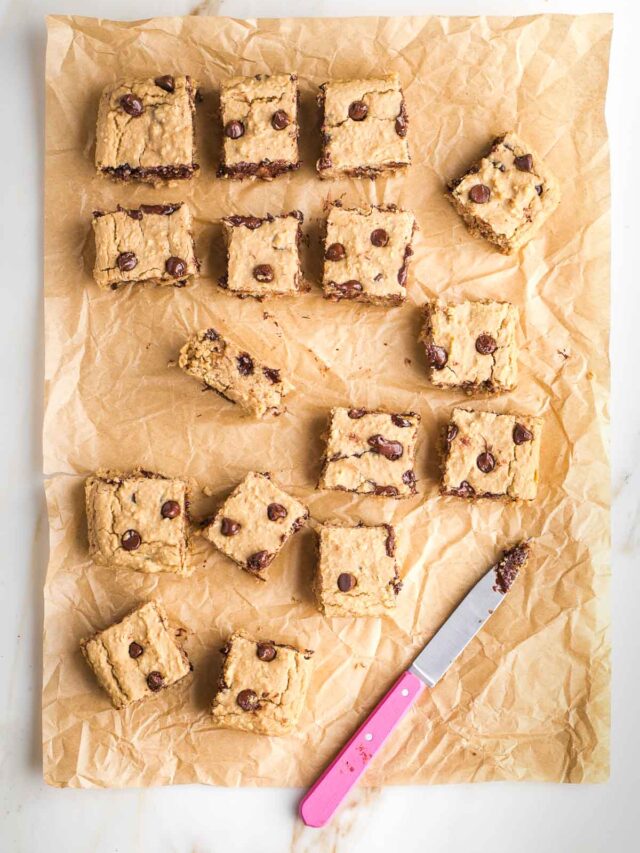 Blondies with chocolate chips on a piece of parchment, cut into squares with a pink knife.