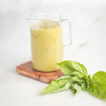Small glass pitcher filled with mustardy yellow colored creamy dressing, with sprigs of basil in the foreground.
