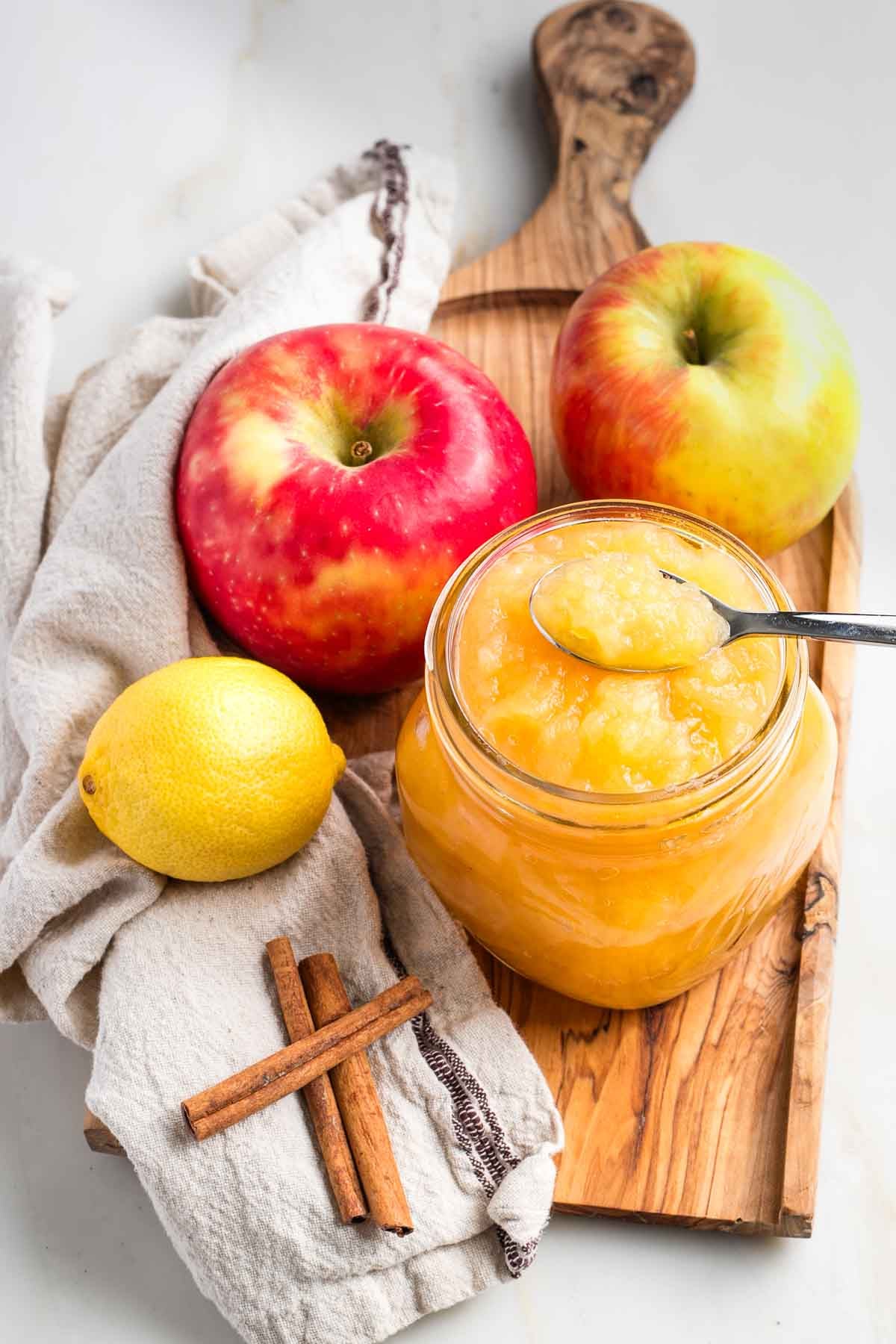 Jar of applesauce with apples, a lemon and cinnamon sticks surrounding.
