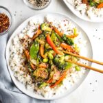 Plate of rice covered with vegetables and teriyaki sauce, sprinkled with sesame seeds.