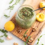 Jar of pesto sprinkled with crushed red pepper, plus lemons, walnuts, arugula and garlic.