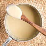 Bechamel sauce clinging to a wooden spoon resting atop a saucepan filled with the white sauce.