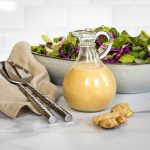 Glass pitcher of salad dressing with salad in the background and ginger root in the foreground.