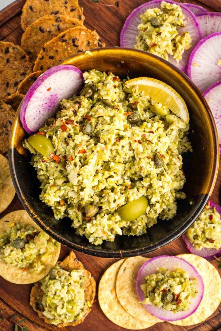 Bowl of green olive tapenade with capers and artichoke hearts, surrounded by crackers and radishes.