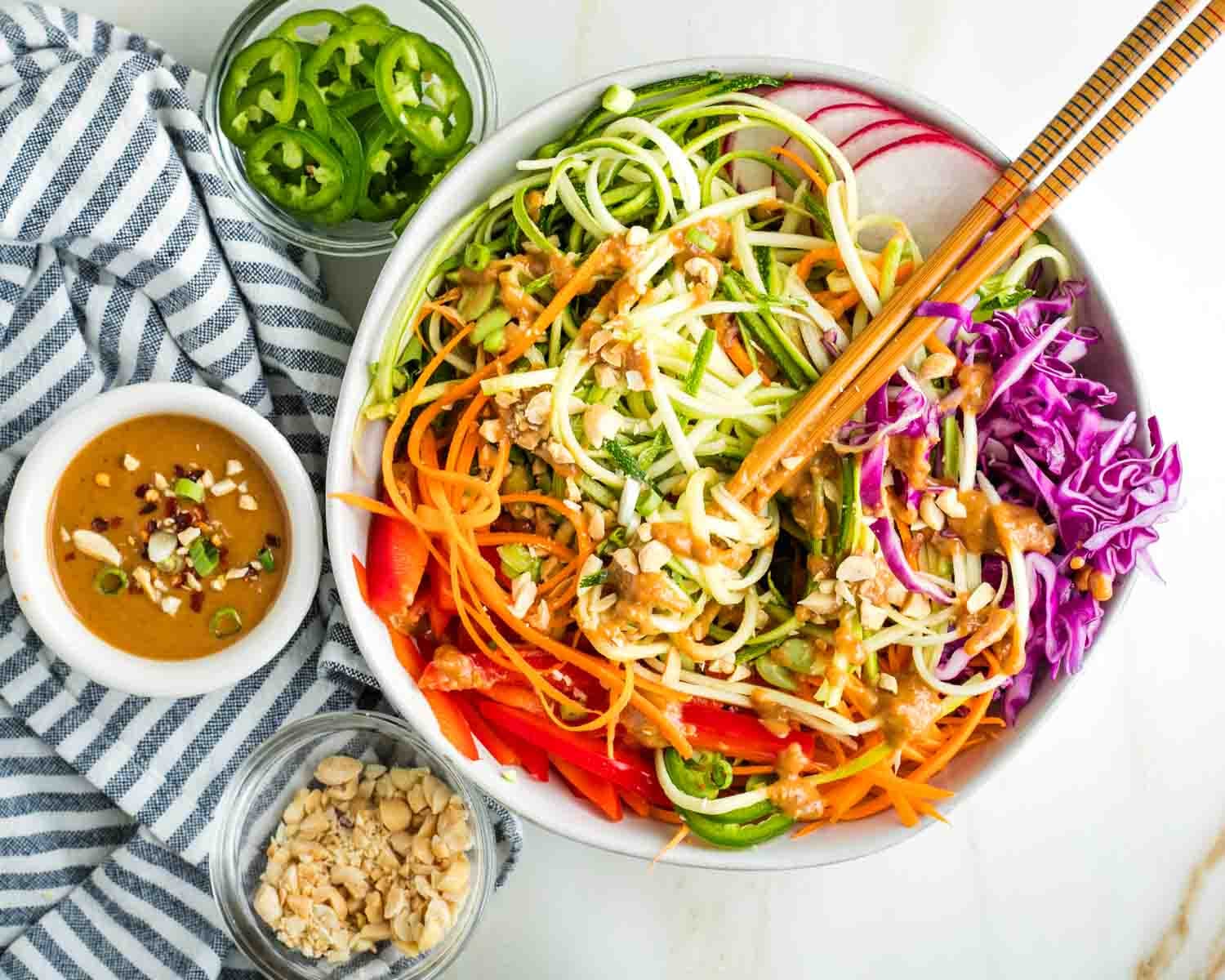 Spring roll bowl with twirled zucchini pasta with chopsticks in a bowl full of vibrant veggies with peanut sauce, chopped peanuts and jalapenos.