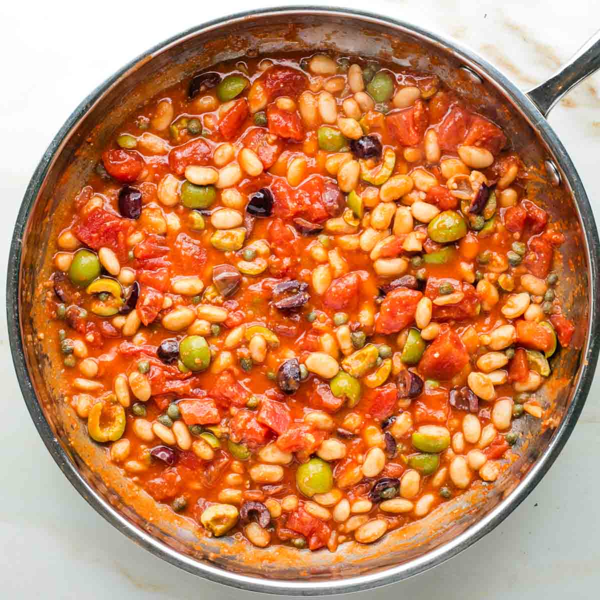 Making puttanesca sauce in a skillet with tomatoes, olives, capers and white beans.