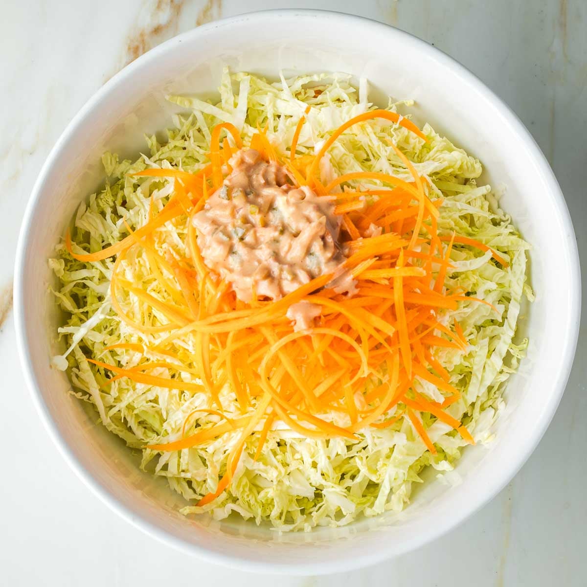 A pile of Russian dressing, chunky with pickles on top of a bowl of shredded cabbage and julienned carrots.