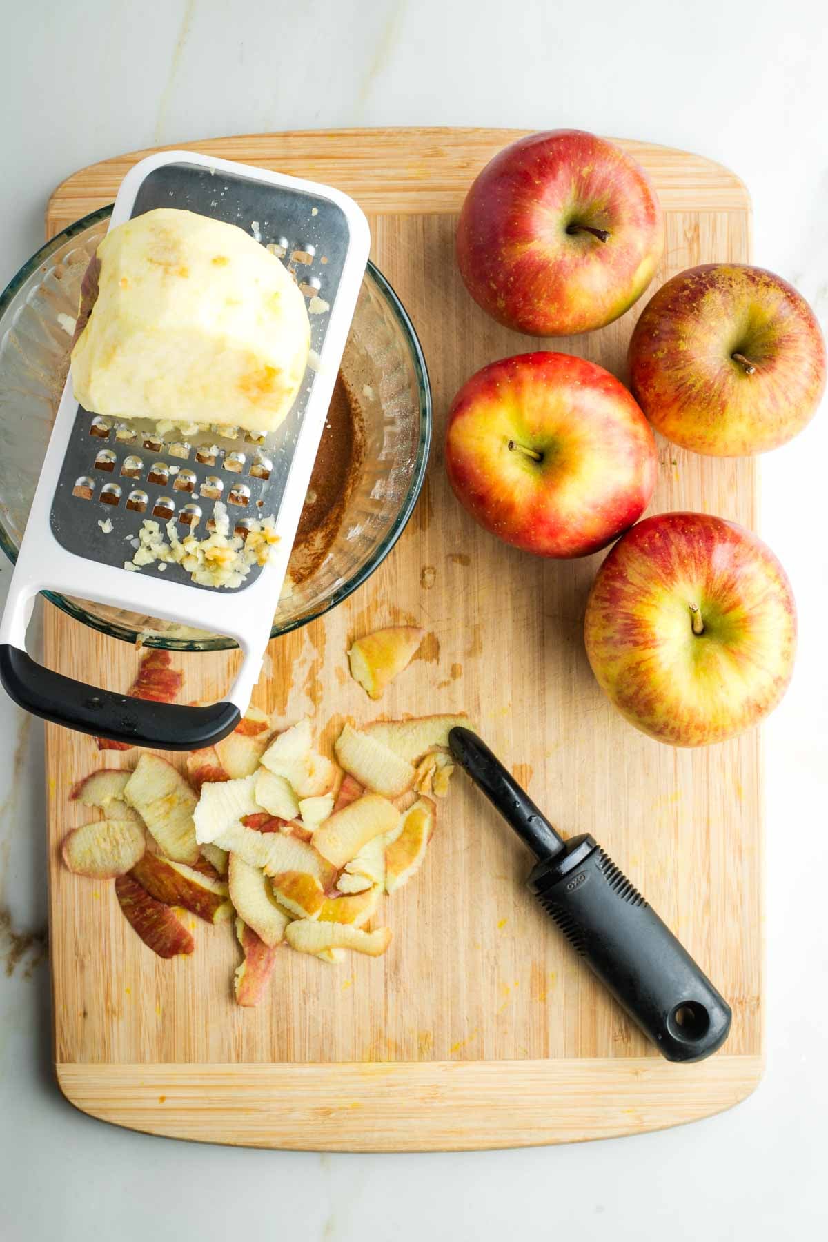 Apples, and apple peels, plus a peeled apple being grated into a glass bowl.