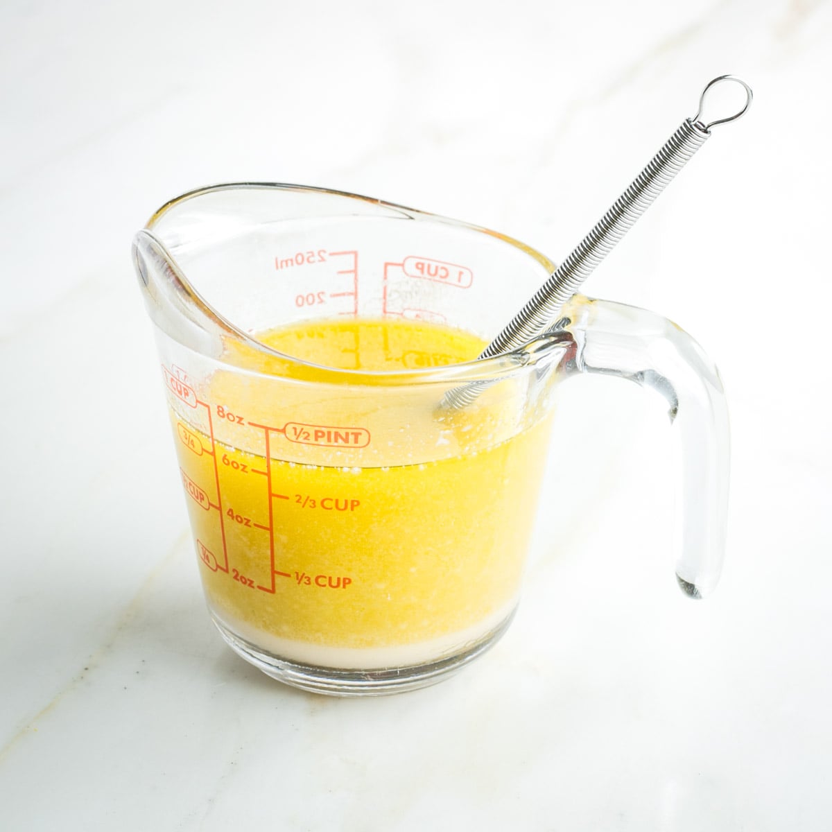 Whisk inside a one cup pyrex measuring cup filled with yellow liquid.