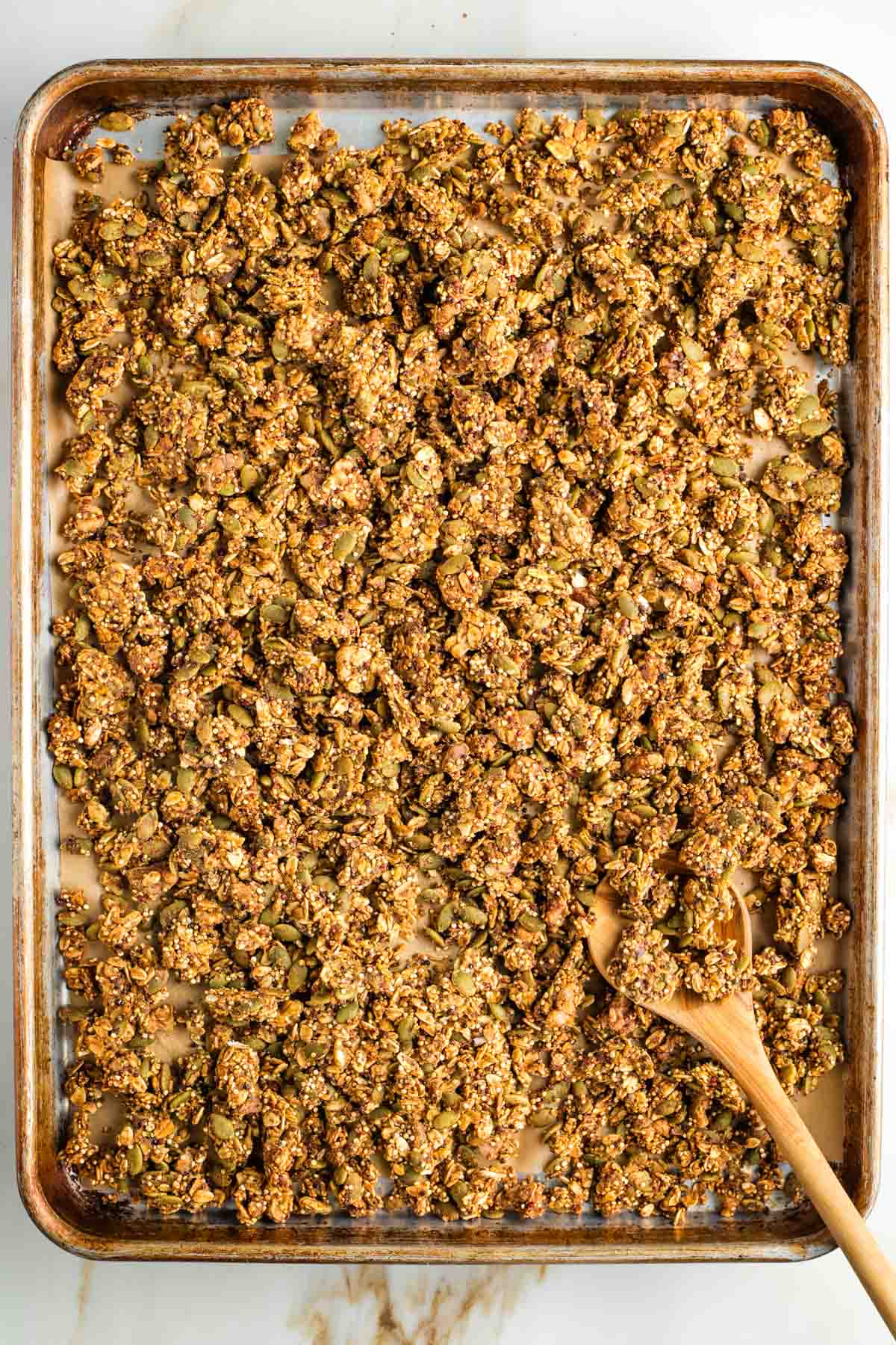 Wooden spoon, pushing around clusters of granola on a large rimmed baking sheet.