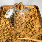 Filling a jar of granola with a wooden spoon from a sheet pan to a mason jar with lid.