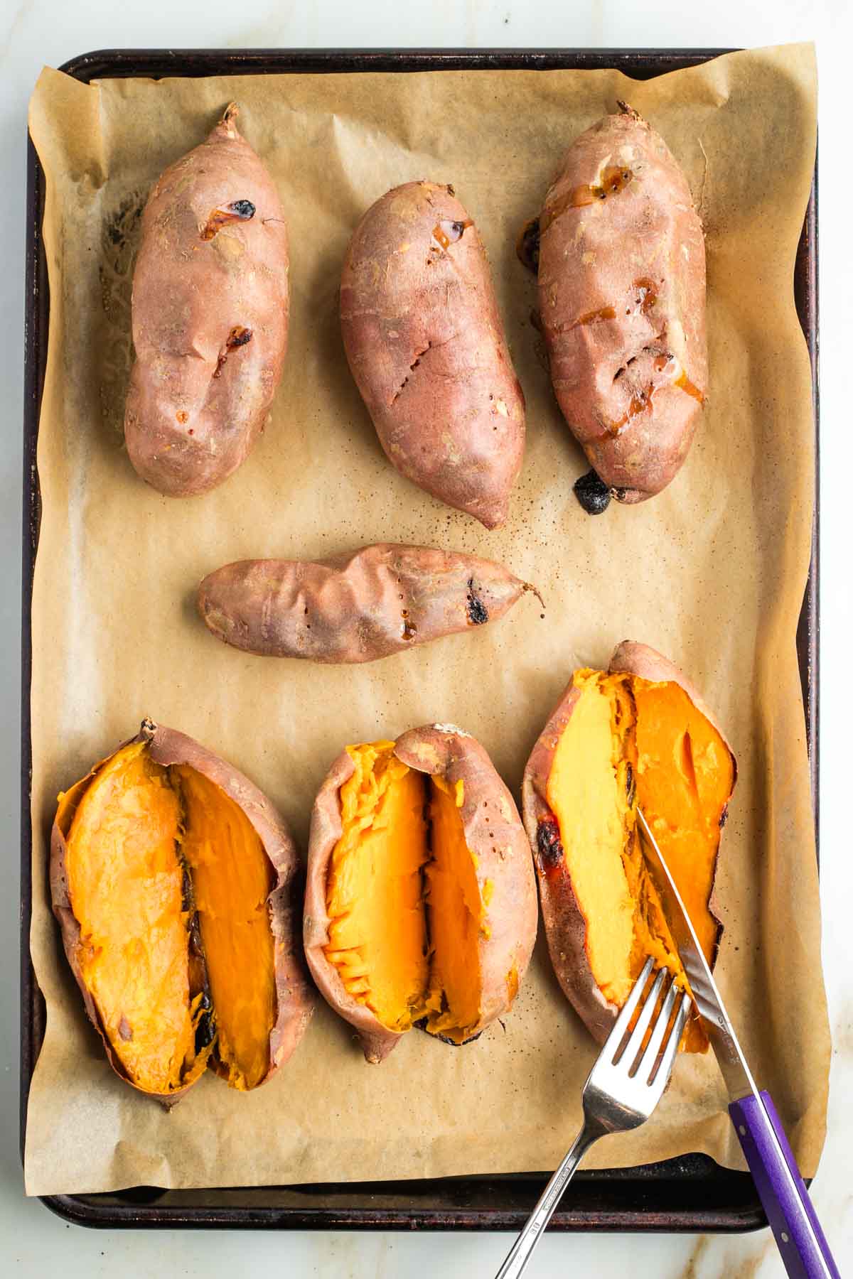 Cutting baked sweet potatoes in half lengthwise, on a tray of roasted whole potatoes. 