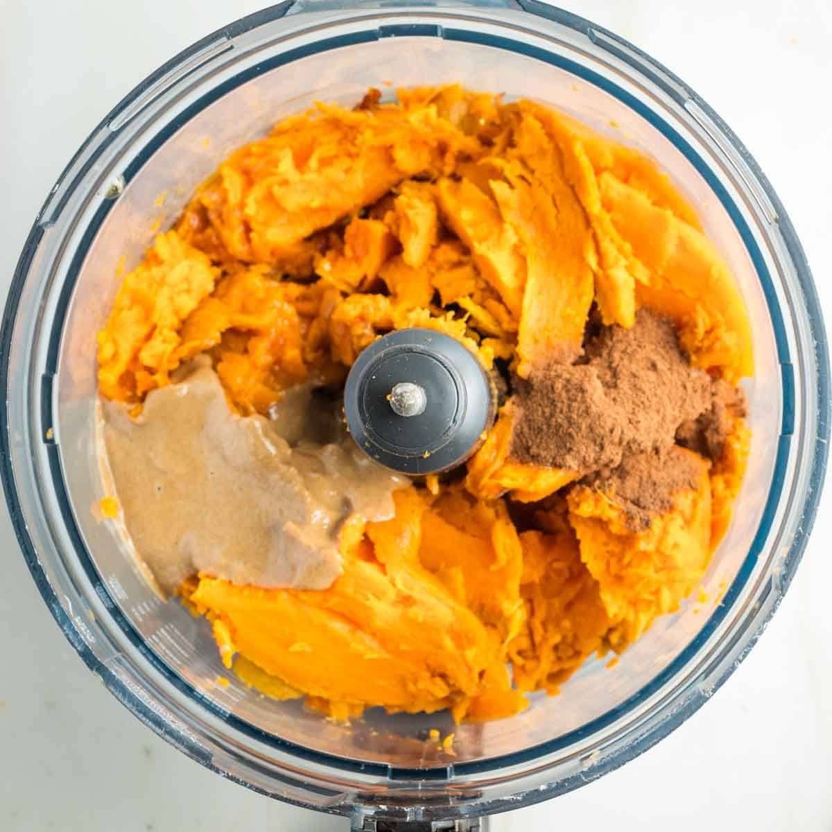 Bowl of food processor with already baked sweet potato pulp, tahini, maple syrup and spices. 