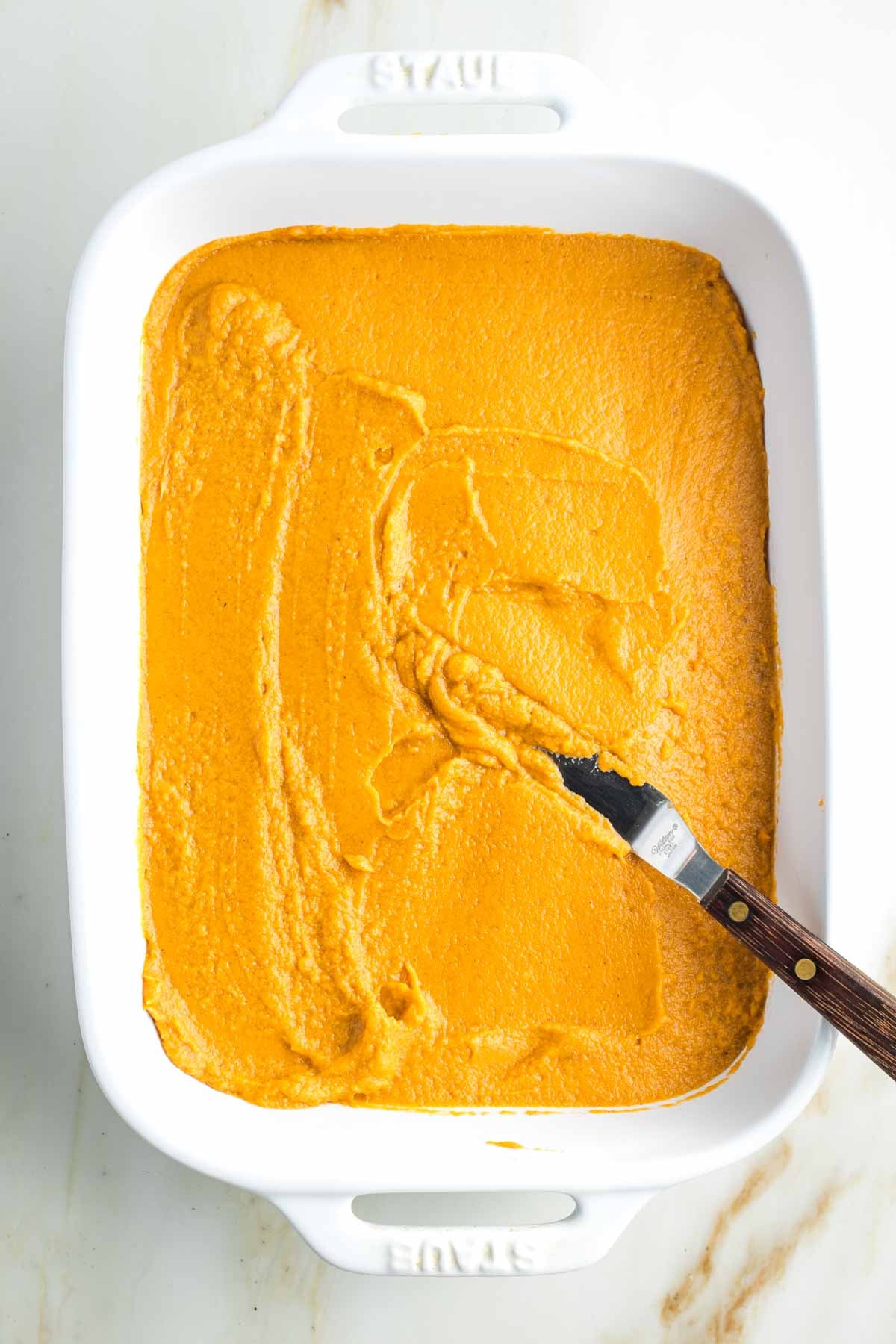 Offset spatula spreading a layer of pureed sweet potatoes into an even layer of a baking dish. 