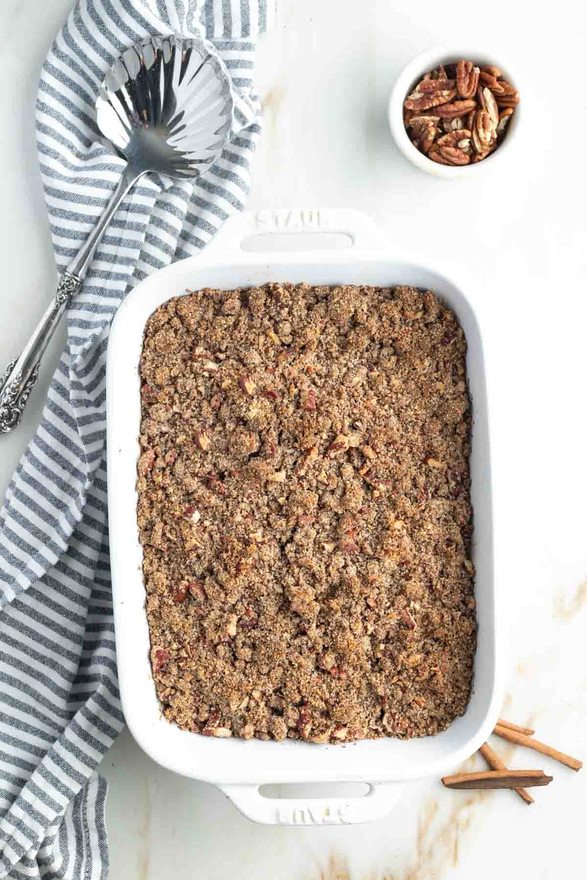 Sweet potato casserole with crumbly pecan topping in a baking dish, styled photo with pecans, cinnamon stick and a serving spoon. 