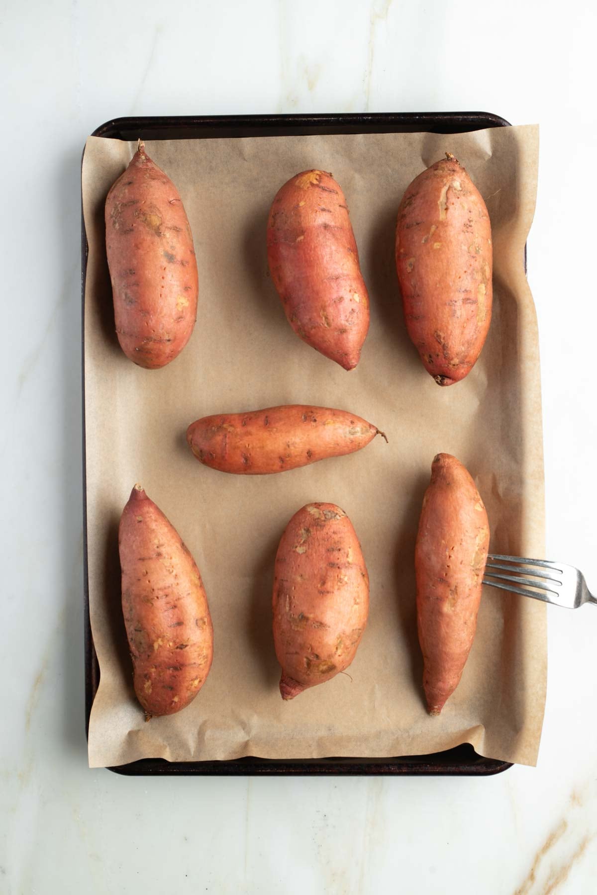 Sweet potatoes, lined up on a parchment lined baking sheet, one being pierced with a fork. Before baking. 