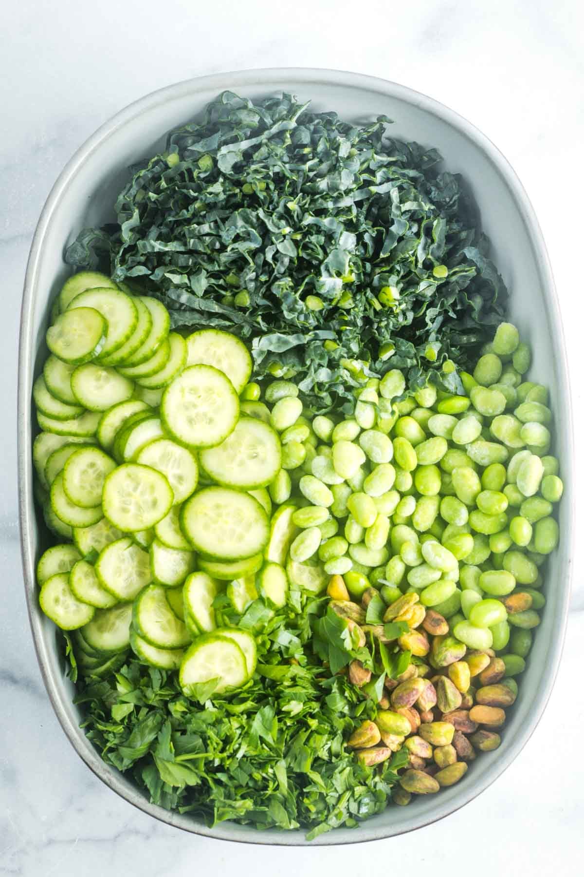 Large oval bowl with sections of julienned kale, thinly sliced cucumber, edamame beans, chopped parsley and pistachios. 