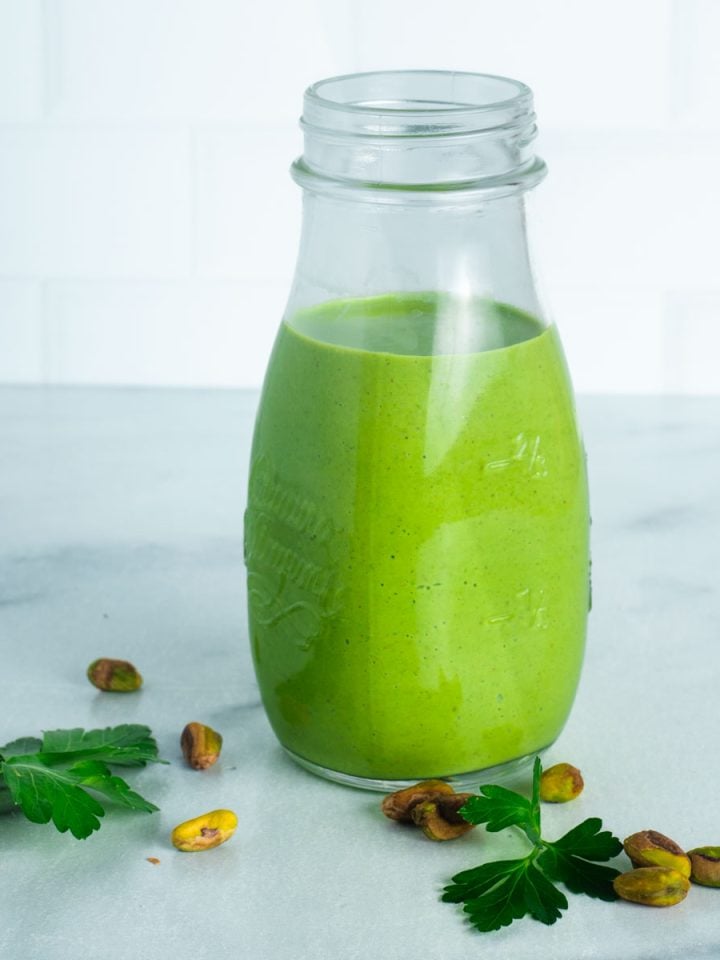Creamy green dressing in a bottle with fresh parsley and pistachios in the foreground.