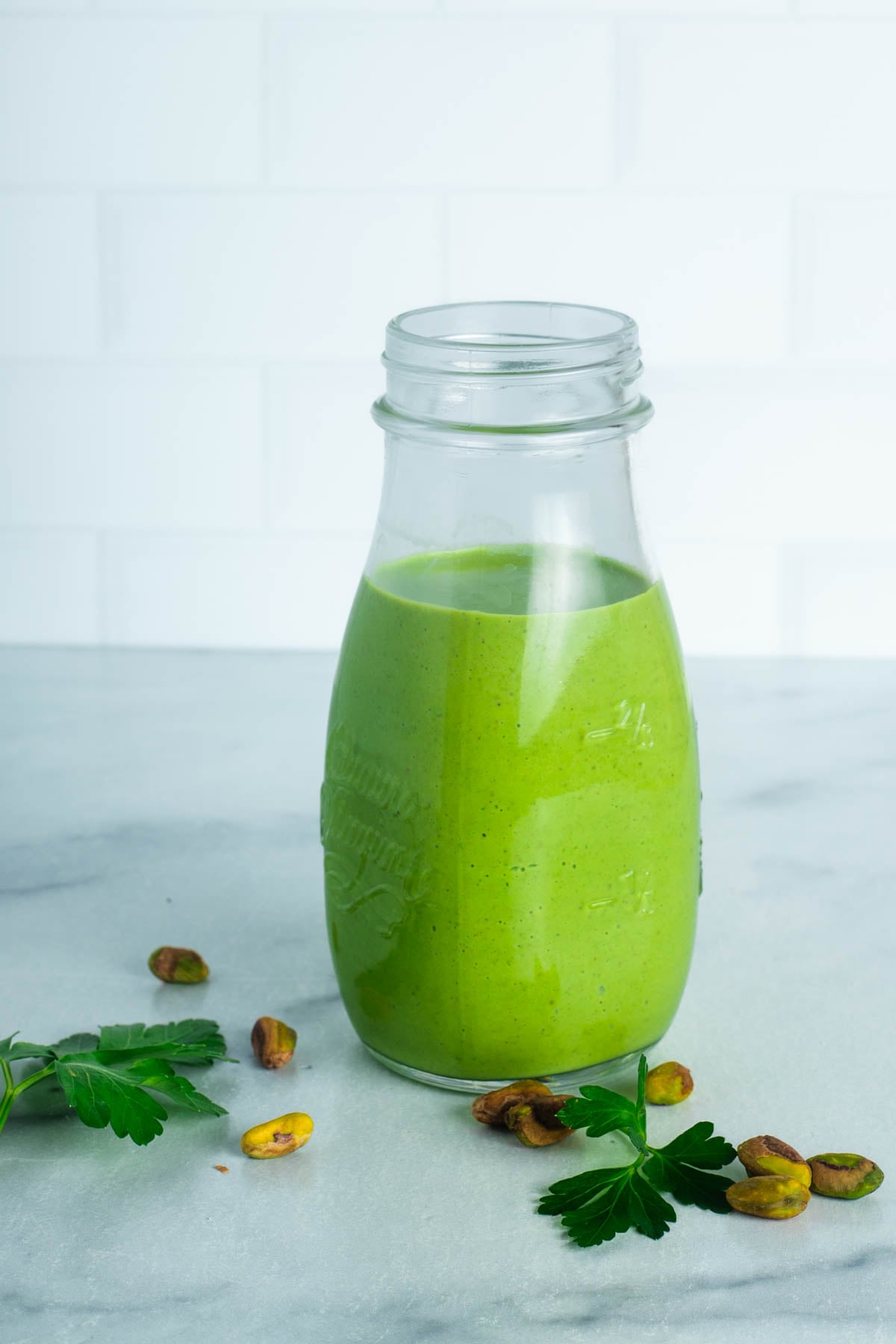 Creamy green dressing in a bottle with parsley leaves and pistachios on the counter. 