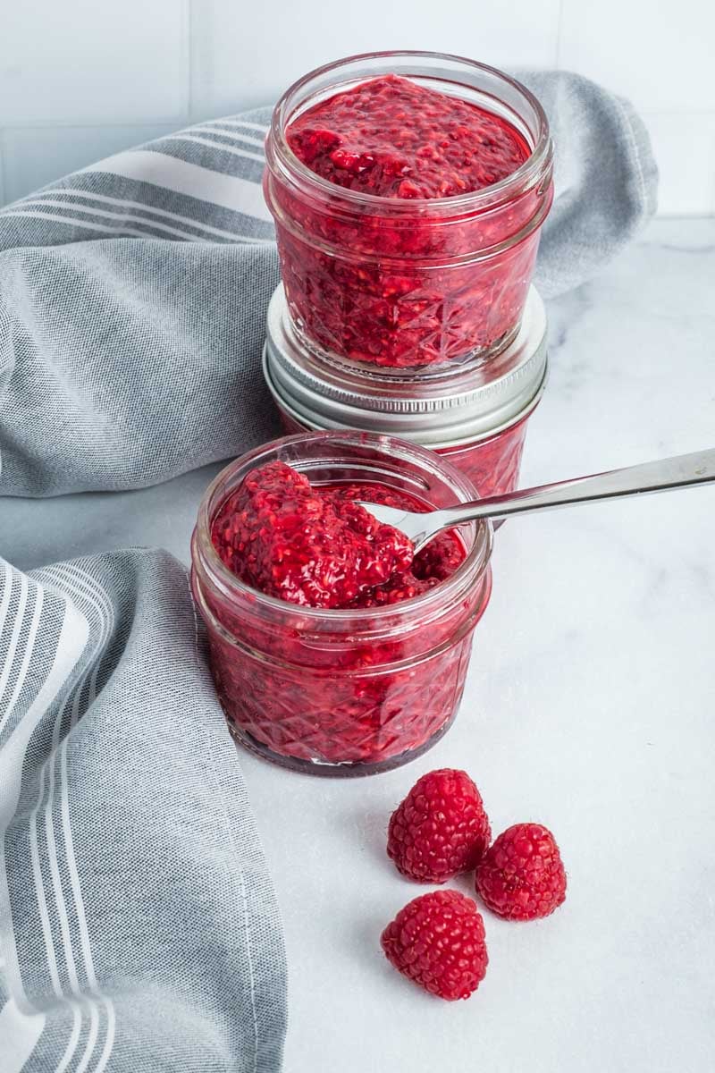 Small mason jars with raspberry chia jam and a few fresh raspberries on the counter.
