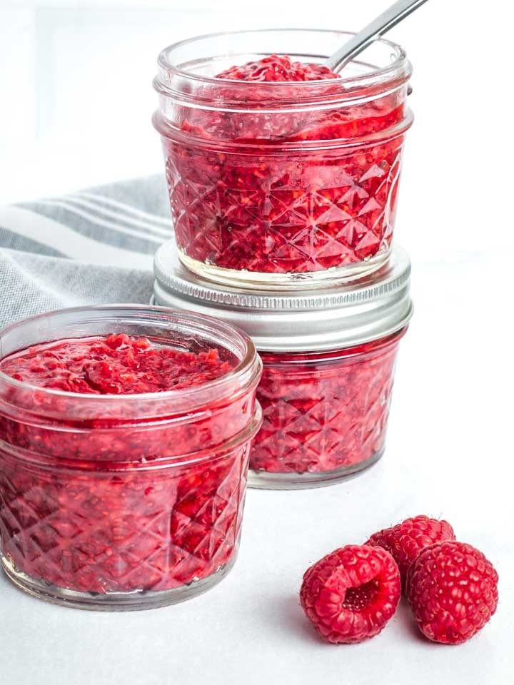 Small jars, filled with raspberry chia jam and a few fresh raspberries on the counter.