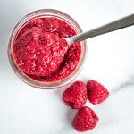 Spoonful of raspberry chia jam on top of the jar, plus three red raspberries on the counter.