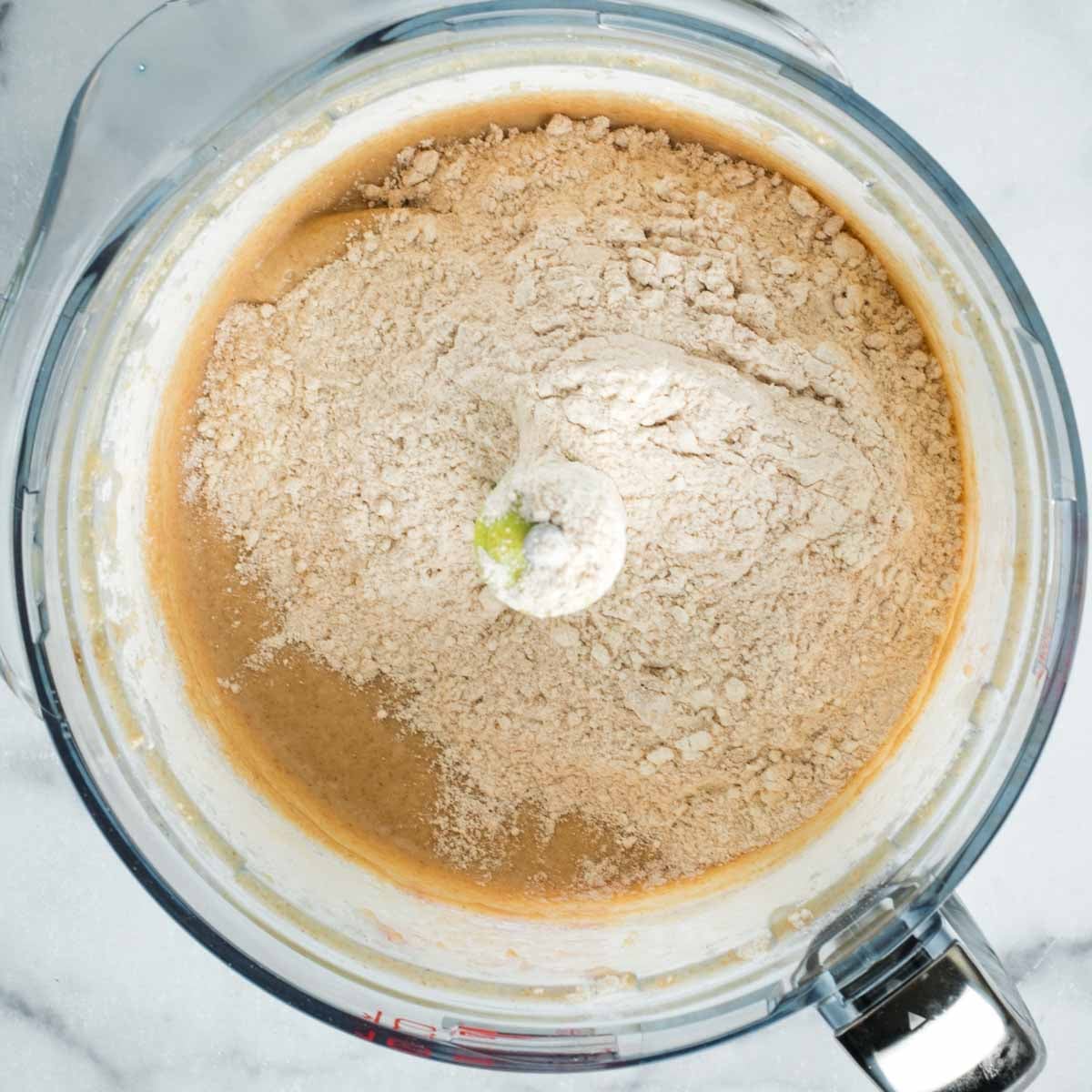 Flour poured on top of sticky donut dough in the bowl of a food processor.