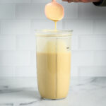 Spoon showing drippy consistency of creamy sauce in a blender cup.