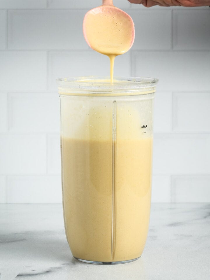 Spoon showing drippy consistency of creamy sauce in a blender cup.
