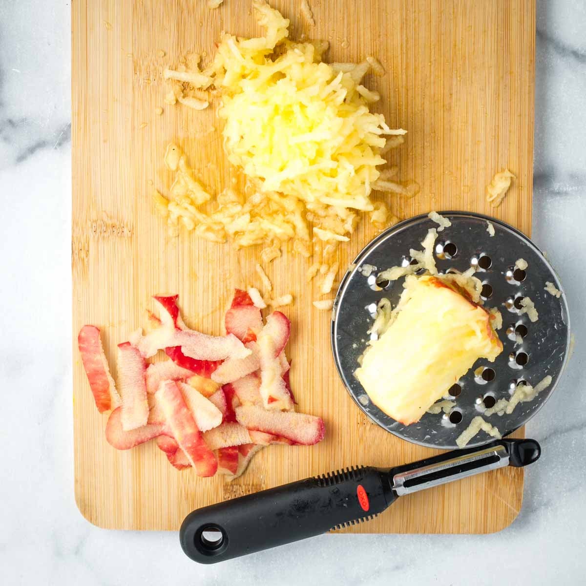 Shredding an apple on a grater with red apple peels on the side of the cutting board with a vegetable peeler. 