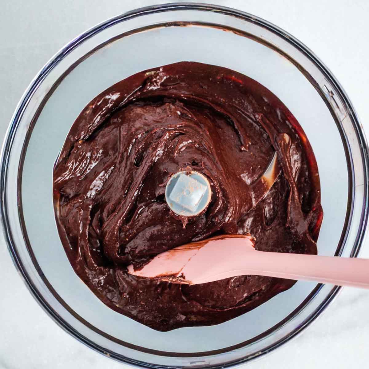 Food processor bowl with pink spatula pushing around chocolate frosting.