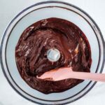 Pink spatula stirring chocolate frosting in the bowl of a food processor.