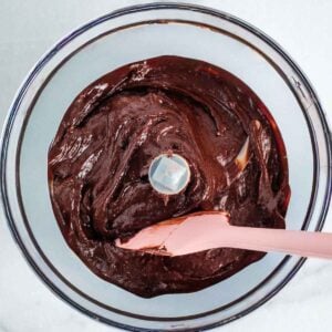 Pink spatula stirring chocolate frosting in the bowl of a food processor.