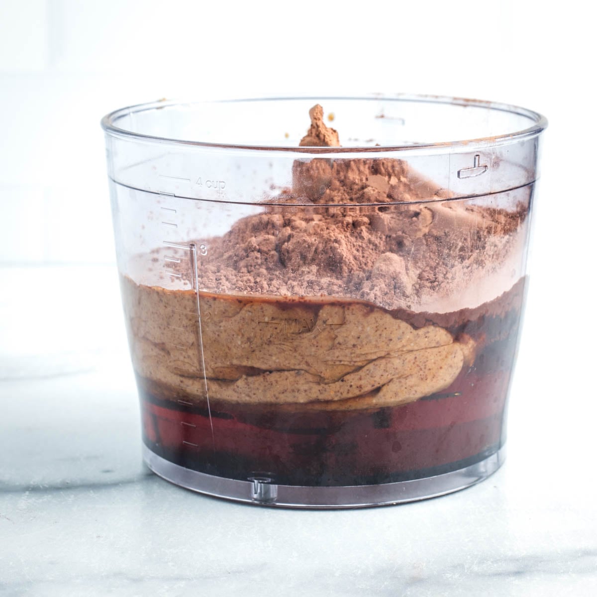 Side view of a food processor bowl with maple syrup, almond butter and cacao powder.