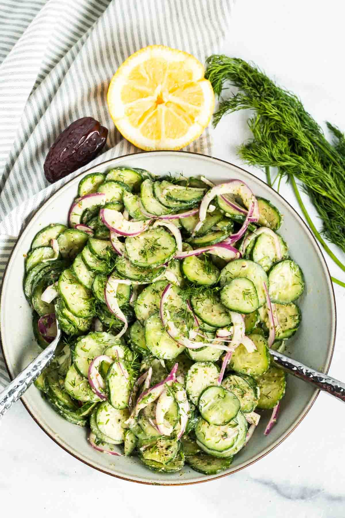 Cucumber, red onion and dill salad with creamy lemon dressing in a bowl with dates, lemon and fresh dill surrounding. 