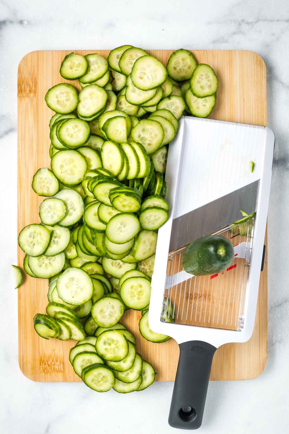 Handheld mandoline slicer on a board with lots of thinly sliced cucumber. 