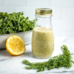 Lemon dill dressing in a bottle with some arugula, fresh dill and half a lemon in the background.