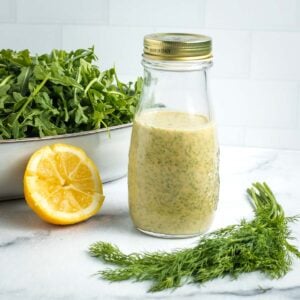 Lemon dill dressing in a bottle with some arugula, fresh dill and half a lemon in the background.