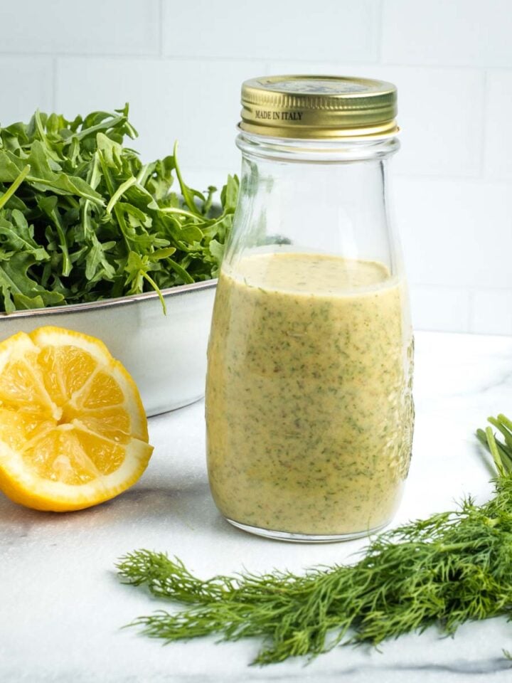 Lemon dill dressing in a bottle with some arugula, fresh dill and half a lemon in the background.