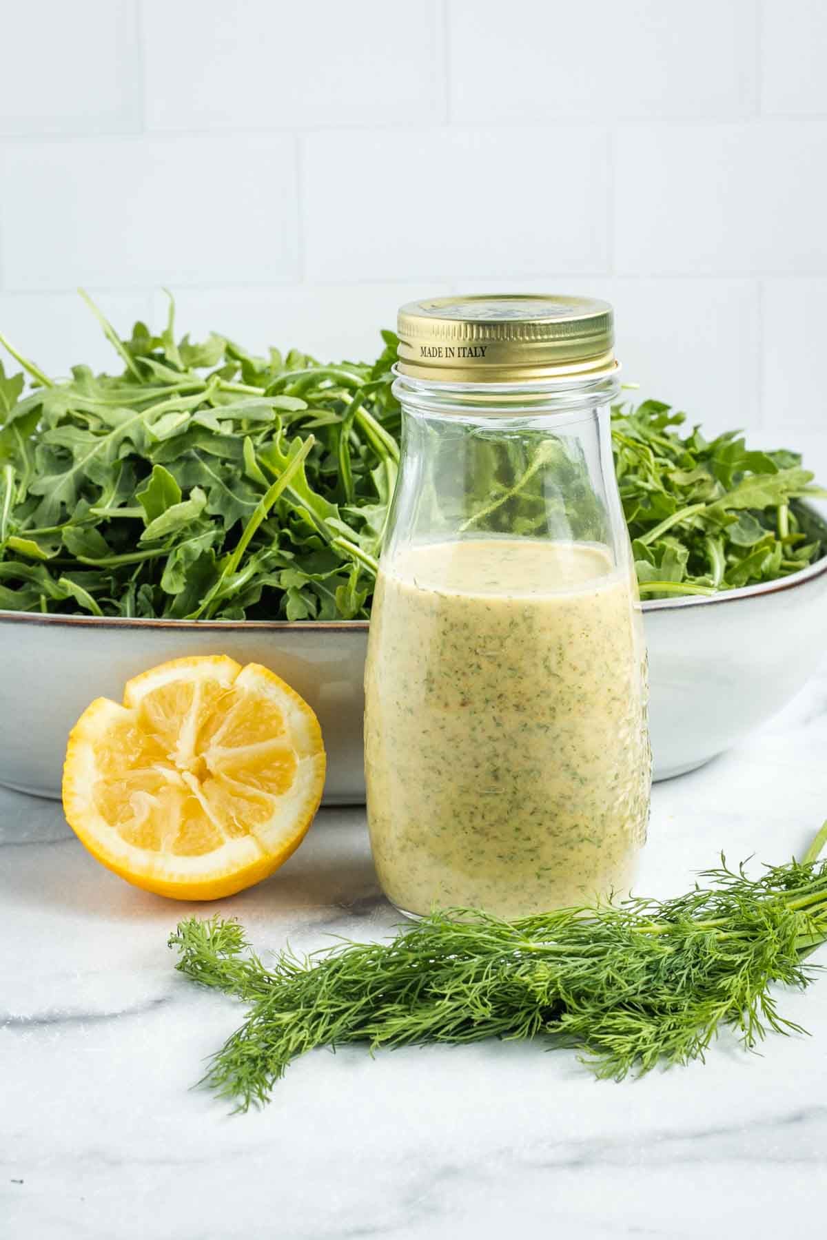 Bottle of creamy dressing in front of a bowl of arugula with half a lemon and some fresh dill. 