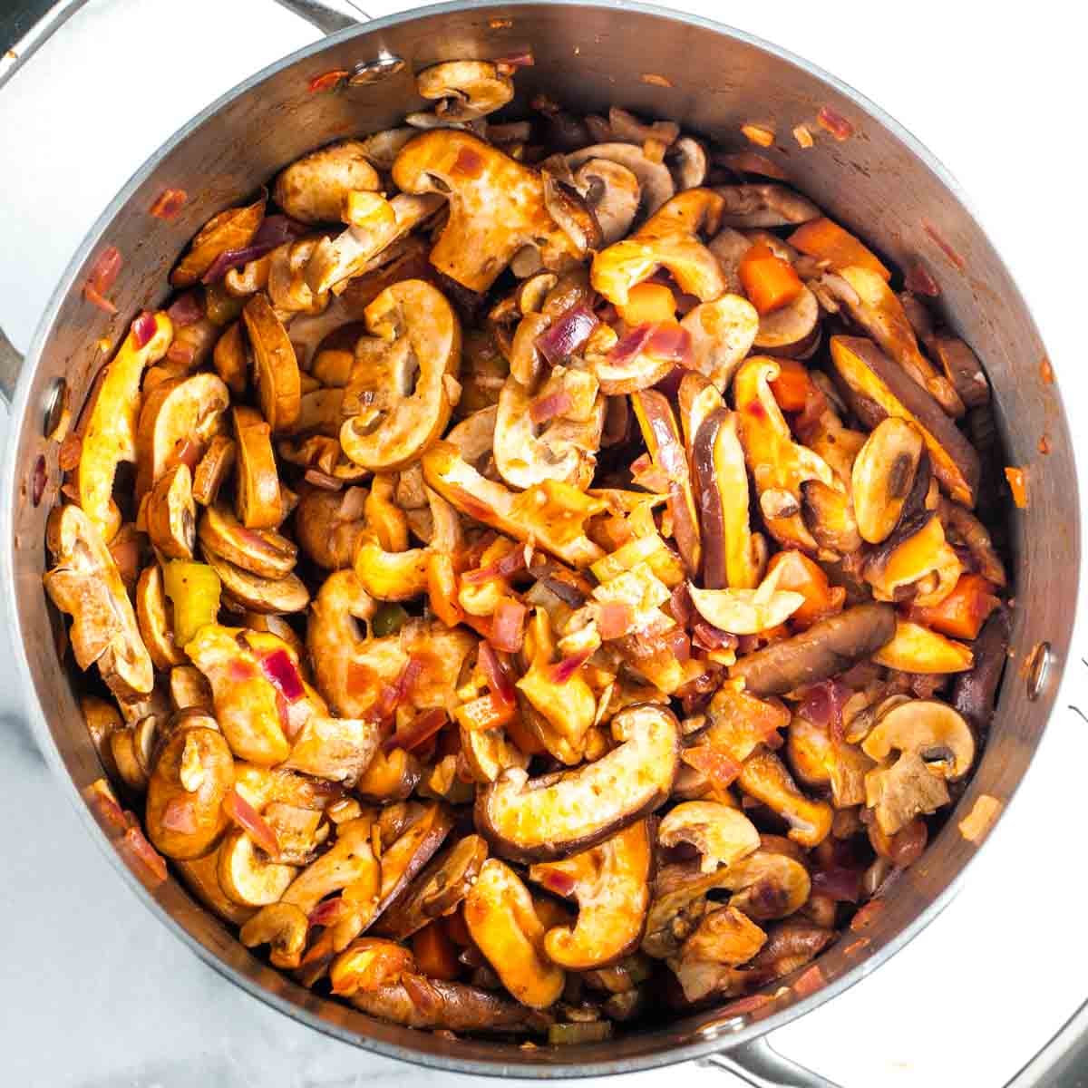 Sliced mushrooms, with diced carrot, onion and celery, sauteed in tomato sauce in a soup pot.