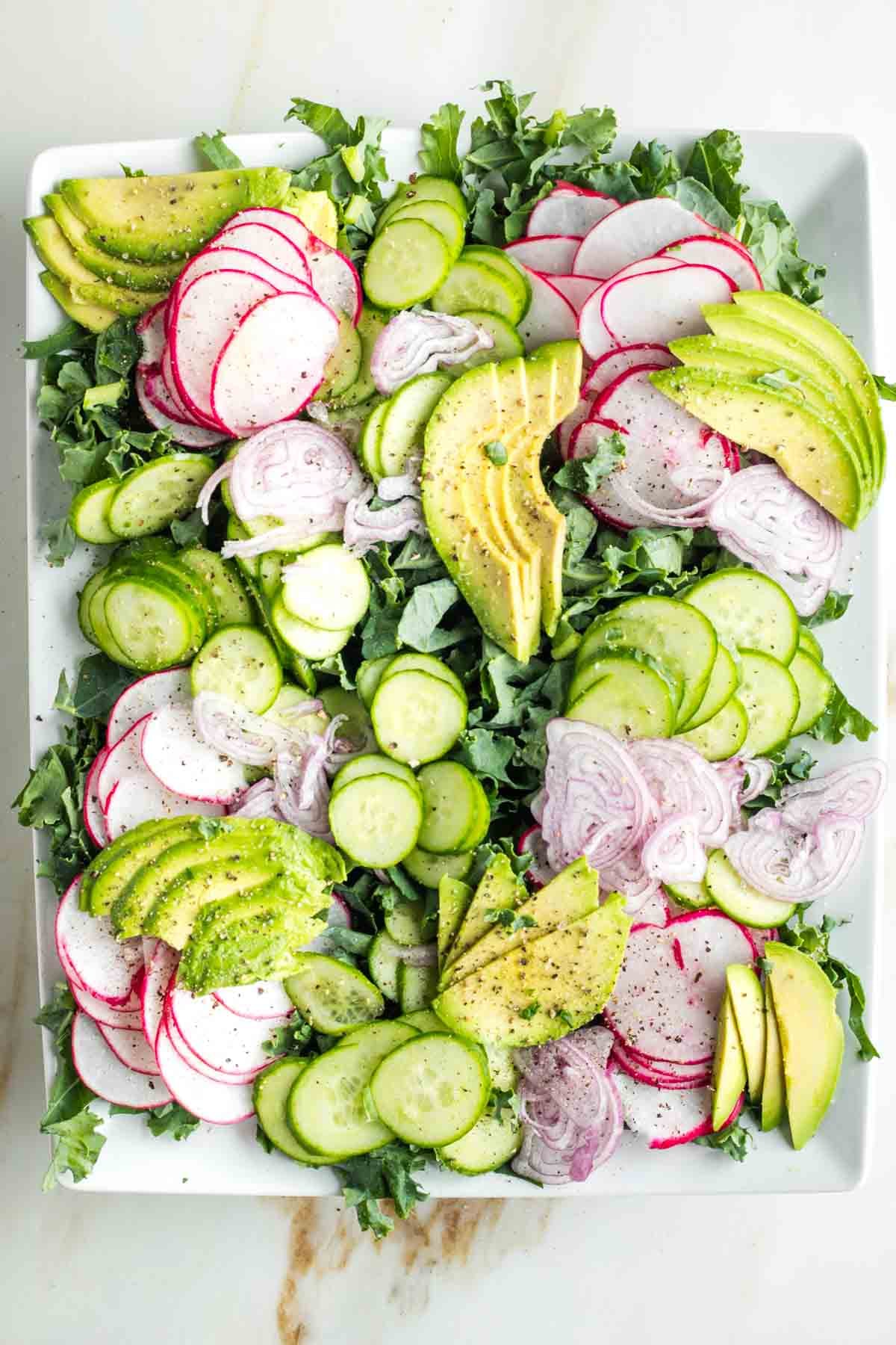 Julienned kale tops with sliced radish, cucumber, avocado and shallot on a platter.
