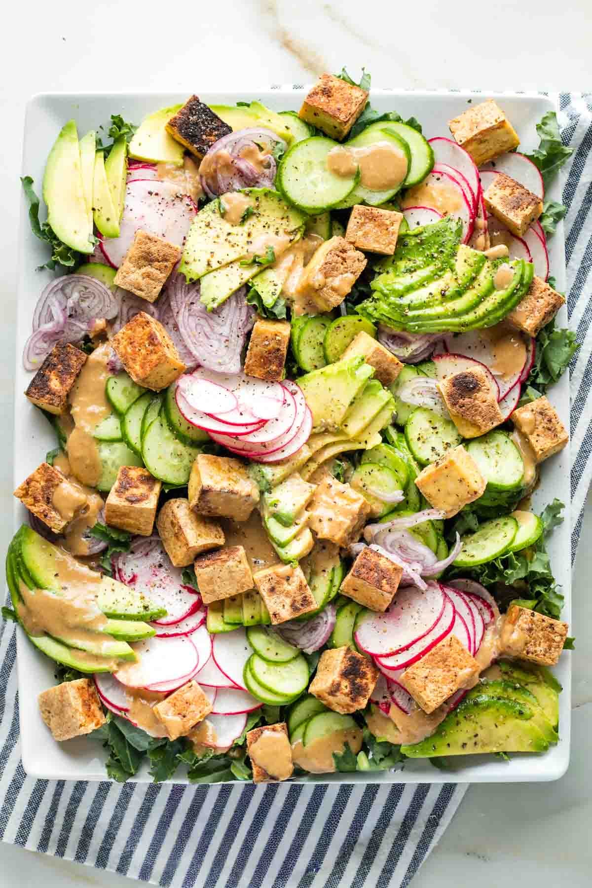 Large platter with slices of cucumber, shallot, radish and avocado on a bed of kale with crisp cubes of tofu, drizzled with sesame ginger dressing.