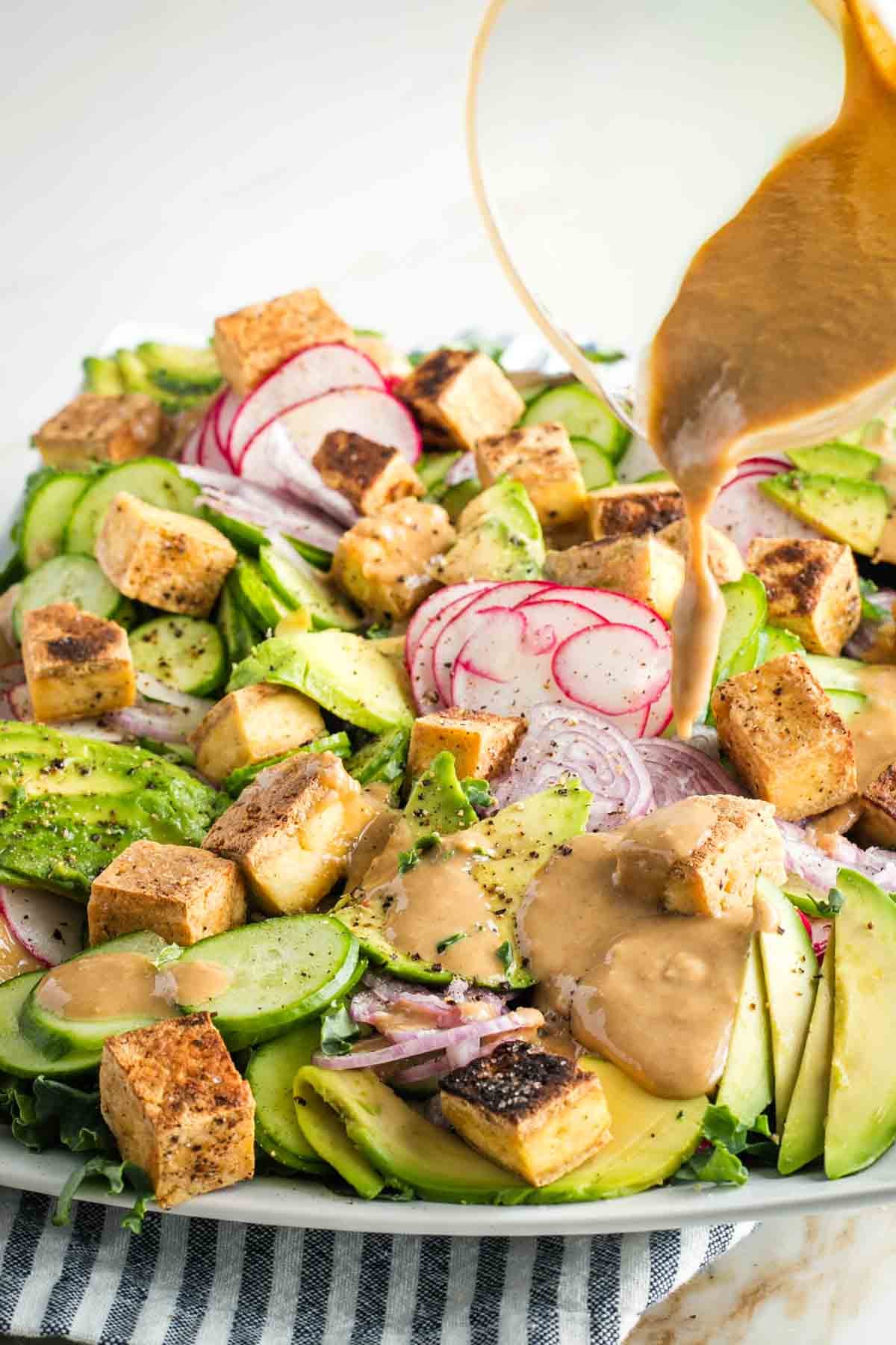 Pouring sesame ginger dressing over a composed salad with slices of avocado, cucumber, radish and shallot plus cubes of tofu.