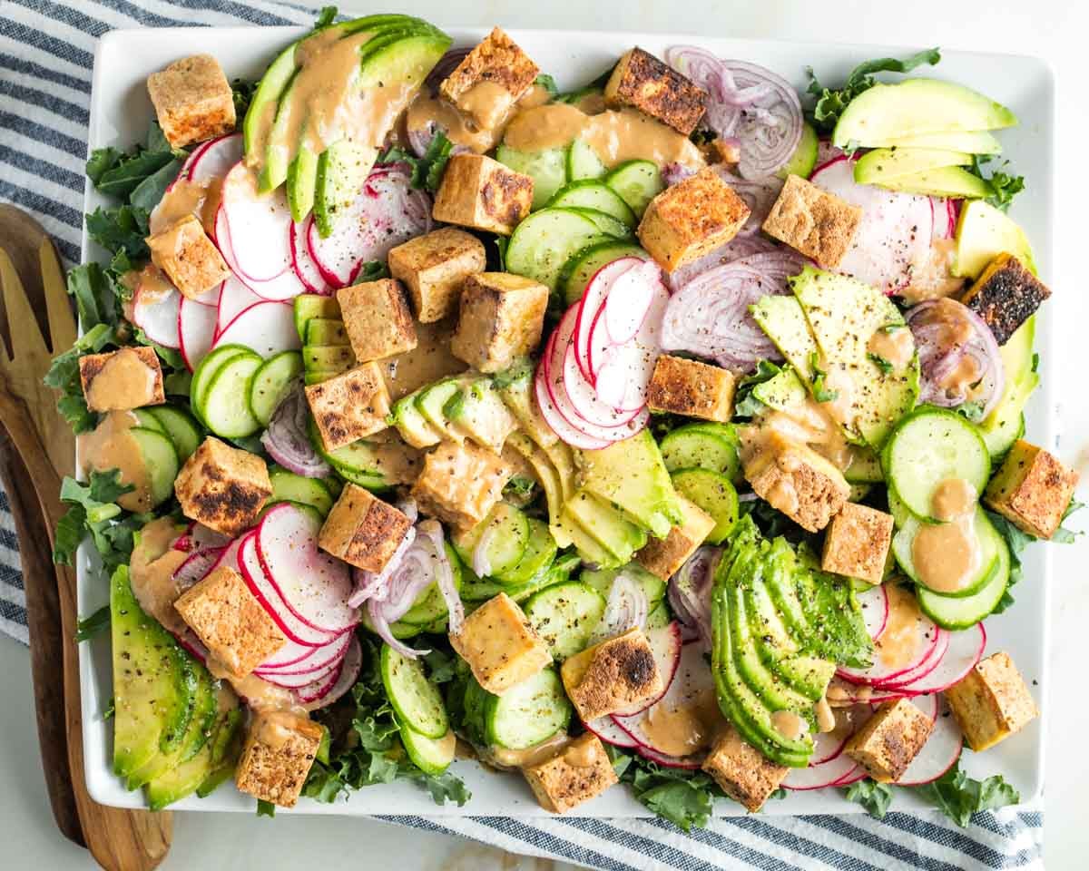 Composed salad with greens, sliced veggies, cubes of tofu, and avocado, drizzled with creamy orangish dressing.